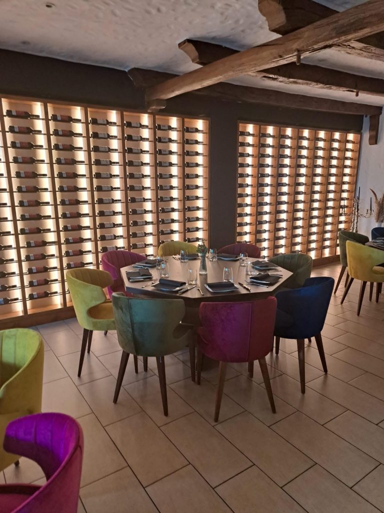 Gastronomie Ausstattung Bunte Stühle um einen Tisch vor einer Wand mit Weinfächern beleuchtet.