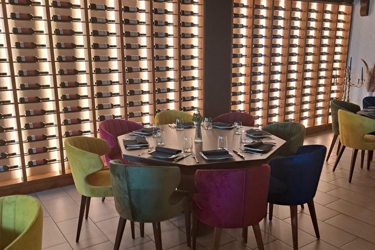 Weinregal mit Stühlen und Tisch Bunte Stühle um einen runden Tisch, umgeben von einer Wand mit Weinregalen.